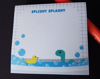 Può includere: Un blocco note bianco con un motivo a griglia e il testo "Splishy Splashy" in alto. Il blocco note presenta un'illustrazione di cartone animato di un'anatra di gomma gialla e un mostro marino verde in una vasca da bagno con acqua blu e bolle.