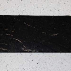 May include: A rectangular black stone serving board with a smooth, polished surface featuring subtle grey and white veining throughout. The board includes a short, integrated handle on one of the shorter sides, creating a flat, elongated shape.