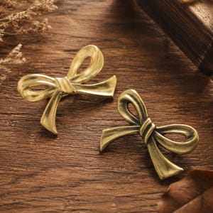 May include: Two metallic bow-shaped pins with a polished gold-tone finish. Each pin features a central knot detail and two flowing ribbon ends, crafted with a smooth, curved surface that reflects light across the bow structure.