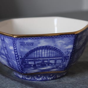 May include: A hexagonal ceramic bowl featuring a blue and white transferware pattern depicting the Tyne Bridge in Newcastle. The bowl has a white interior and a gold-colored rim along the top edge and base, with the text Tyne Bridge Newcastle.