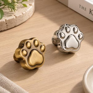 May include: Two metal cabinet knobs shaped like animal paw prints. One knob features a brushed antique brass finish, while the other has a polished silver-tone finish. Each knob displays a central pad and four smaller toe pads.