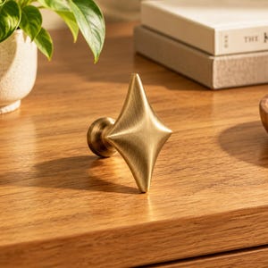 May include: A brass-toned metal cabinet knob featuring a four-pointed star or diamond shape with a brushed finish. The knob has a central circular base that connects to the elongated, faceted geometric front piece, creating a distinct point.