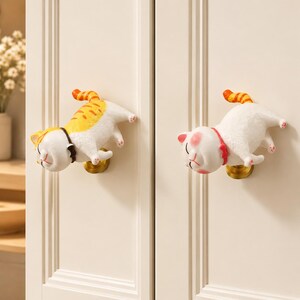 May include: Two white and orange cat-shaped cabinet knobs mounted on cream-colored doors. Each knob features a sculpted cat with a striped tail, a small collar, and pink paw pads, attached to a circular gold-toned metal base for easy gripping.