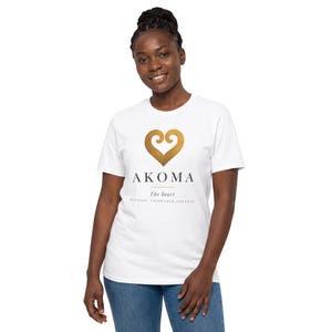 Puede incluir: Una camiseta blanca de manga corta con un símbolo dorado en forma de corazón sobre el texto AKOMA, seguido de las palabras The heart y PATIENCE, TOLERANCE AND LOVE impresas en negro en el centro del pecho.