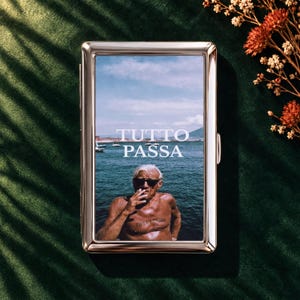 May include: A rectangular silver-toned metal cigarette case featuring a glossy photograph of a sun-tanned person wearing sunglasses by the sea, with the white text TUTTO PASSA printed across the center of the image.