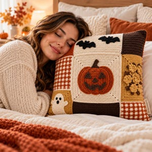 Puede incluir: Un cojín de ganchillo cuadrado con un motivo central de calabaza, un fantasma, murciélagos y detalles florales en un diseño de patchwork. El cojín está elaborado con hilo de color crema, naranja, marrón y amarillo.