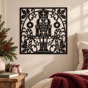 May include: A square black metal wall art piece featuring a central nutcracker figure surrounded by decorative holly leaves, candy canes, stars, and circular ornaments within a symmetrical, intricate cutout frame design.
