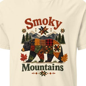 May include: A cream-colored short-sleeve t-shirt featuring a graphic of a bear with a patchwork quilt pattern. Above the bear is the text "Smoky" and below is "Mountains" in dark green. The design includes pine trees, mountains, and autumn leaves.