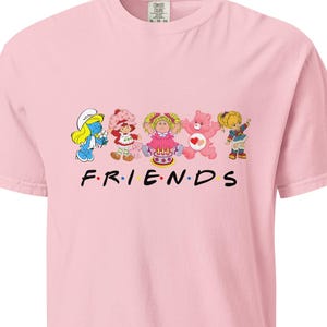 May include: A light pink t-shirt featuring cartoon characters including a Smurf, Strawberry Shortcake, and Care Bears. The word "FRIENDS" is printed in black block letters below the characters. The shirt is made of a soft material.
