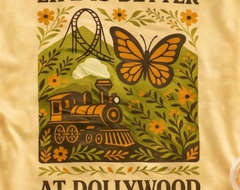 May include: A pale yellow short-sleeve t-shirt featuring a graphic print with the text 'LIFE IS BETTER AT DOLLYWOOD'. The design includes a steam locomotive, a roller coaster, a large monarch butterfly, and various flowers in shades of orange and green.