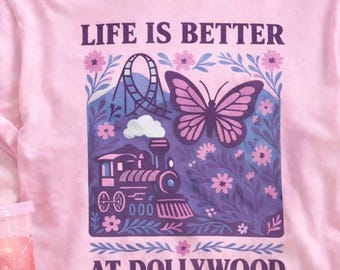 May include: A light pink t-shirt featuring a graphic print with the text "LIFE IS BETTER AT DOLLYWOOD". The design includes a purple butterfly, a steam train, a roller coaster, and floral elements in shades of purple, blue, and pink.