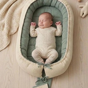 Könnte beinhalten: Ein Baby liegt in einem ovalen Babynest mit einer cremefarbenen, waffelstrukturierten Außenseite und einem salbeigrünen Innenbereich. Das Nest verfügt über ein salbeigrünes Stoffband zum Binden am unteren Ende. Das Baby trägt einen cremefarbenen Strickbody.