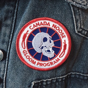 May include: A round patch with the words "CANADA NOOSE GLOOM PROGRAM" encircling a white skull on a blue background. The patch is bordered by a red and white design and is sewn onto a denim jacket.