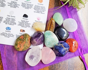 May include: A purple pouch containing a set of 12 tumbled gemstones, including amethyst, aventurine, carnelian, clear quartz, obsidian, rose quartz, sodalite, tiger's eye, and more. The pouch is open, revealing the gemstones. A card with the names and meanings of the gemstones is visible in the background.