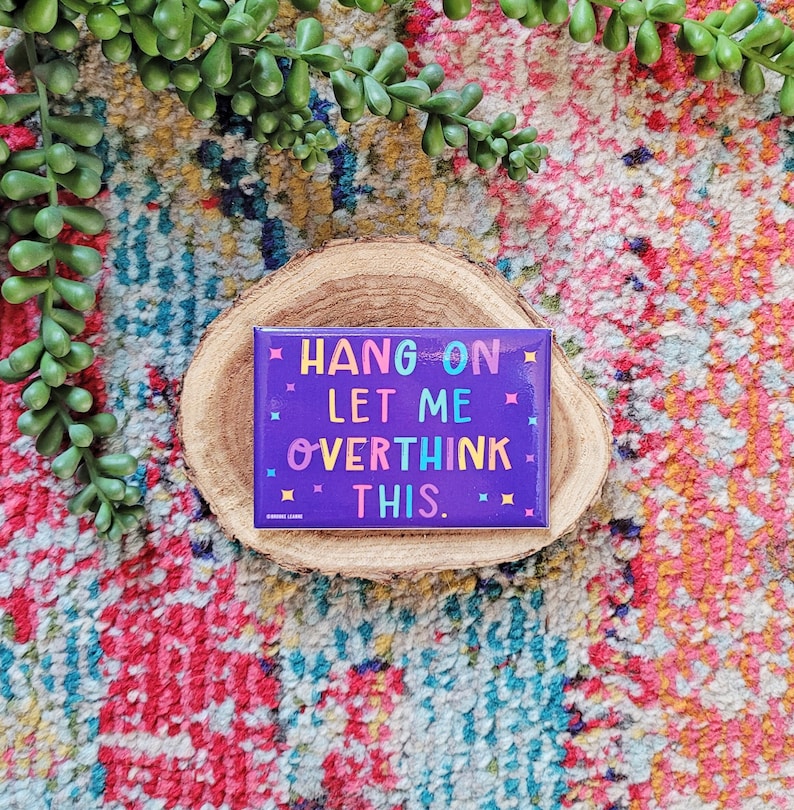 Op de afbeelding: Paarse vierkante magneet met de tekst "HANG ON LET ME OVERTHINK THIS." in kleurrijke letters op een houten achtergrond.