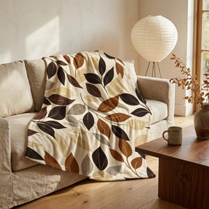 May include: A soft throw blanket featuring a repeating pattern of stylized leaves in shades of dark brown, light brown, tan, and cream. The blanket is draped over a neutral-colored sofa, showcasing the detailed botanical print across its surface.