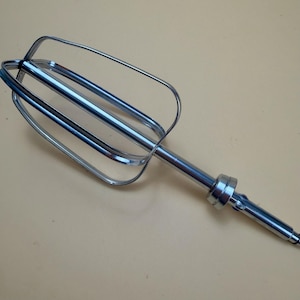 May include: A stainless steel hand mixer attachment featuring a central rod and four curved wire loops that form a cage-like whisking head. The metal surface is reflective and smooth, with a cylindrical connector at the base of the whisking wires.