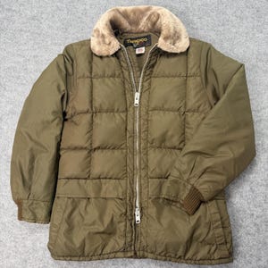 Peut inclure: Une veste matelassée vert olive avec un col en fausse fourrure beige et une fermeture éclair argentée sur toute la longueur. La veste présente une étiquette de marque Tempco au col, des manches longues et des poignets côtelés.