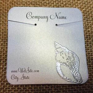 May include: Silver jewelry display card with a seashell design. The card has holes for hanging jewelry and includes the text "Company Name", "www.WebSite.com", and "City, State".