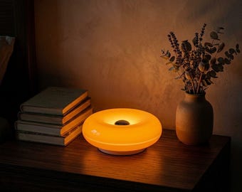 Könnte beinhalten: Eine runde, donut-förmige Lampe, die ein warmes, leuchtendes orangefarbenes Licht ausstrahlt. Die Lampe hat eine glatte, kreisförmige Oberfläche mit einer dunklen Mitte. Sie steht neben einem Stapel aus vier gebundenen Büchern und einer Keramikvase.