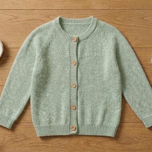Könnte beinhalten: Eine hellgrüne Strickjacke mit langen Ärmeln, geripptem Rundhalsausschnitt und fünf braunen Knöpfen auf der Vorderseite. Eine kleine weiße Schale mit vier zusätzlichen Knöpfen und ein weißes Maßband liegen neben dem Kleidungsstück.