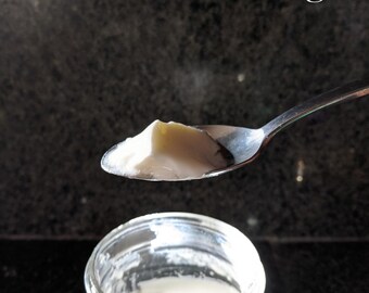 May include: A silver metal spoon holds a portion of thick, off-white sourdough starter above an open glass jar containing the same substance. The text SanFranSourdough is visible at the top and bottom right of the image.