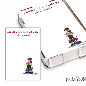 May include: A white notepad with red apple accents and the text "From the Desk of Miss Amanda". The notepad is in a clear plastic holder. A cartoon illustration of a girl sitting on a stack of books is on the notepad.