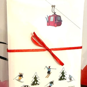 May include: A set of white greeting cards tied with a red ribbon. The cards feature illustrations of skiers, pine trees, and a ski lift. The ski lift is a pink gondola. The artist's signature is in the bottom right corner.