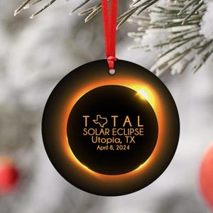 May include: A round, black Christmas ornament with a gold ring around the edge. The ring has a silhouette of the state of Texas inside of it. The text "TOTAL SOLAR ECLIPSE Utopia, TX April 8, 2024" is printed in gold inside the ring.