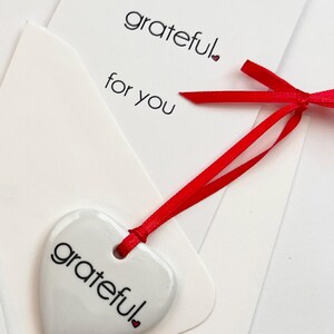 May include: A white heart-shaped ornament with the word "grateful" printed in black, tied with a red ribbon to a white card. The card also has the words "grateful. for you" printed in black. The background is white.