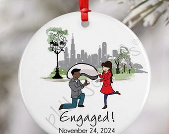 May include: White round Christmas ornament with a cartoon illustration of a couple getting engaged in front of a city skyline. The text "Engaged! November 24, 2024" is printed on the ornament.