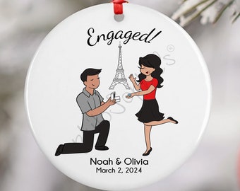 May include: White round ornament with a cartoon illustration of a couple getting engaged in front of the Eiffel Tower. The text "Engaged!" is at the top of the ornament. The text "Noah & Olivia March 2, 2024" is at the bottom of the ornament.