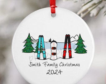 Peut inclure: Une décoration ronde en céramique blanche avec un ruban rouge, illustrée de deux sapins verts, d'une paire de skis bleu clair, d'un snowboard blanc et d'une paire de skis rouges. Le texte indique Smith Family Christmas 2024.