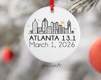 May include: Round white ceramic ornament with a red ribbon. It displays a black and white skyline illustration and the text "ATLANTA 13.1 March 1, 2026". The ornament has a small logo at the bottom.