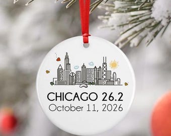 May include: A round white ceramic ornament hanging from a red ribbon. The front features a black line art illustration of the Chicago skyline with small colored accents, the text CHICAGO 26.2, and the date October 11, 2026, printed in black.