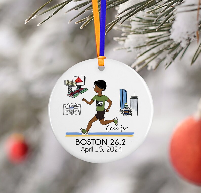 May include: White round ceramic ornament featuring a cartoon illustration of a runner, a cityscape, and the text "BOSTON 26.2 April 15, 2024". The runner is wearing a green shirt with the number "2024" on it and black shorts. The text "Jennifer" is written below the runner.