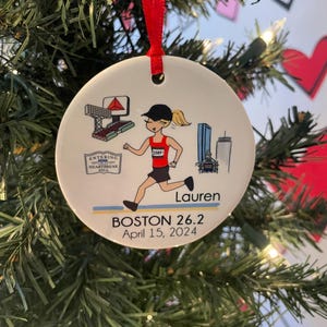 May include: White ceramic bauble featuring a cartoon illustration of a runner wearing a red top and a bib with the number 2589. The bauble also includes a cityscape, a sign that reads "Entering the Heartbreak Hill", and the text "Boston 26.2 April 15, 2024" and "Lauren".