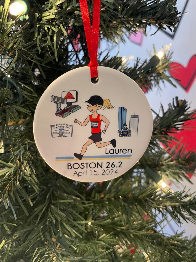 May include: White ceramic bauble featuring a cartoon illustration of a runner wearing a red top and a bib with the number 2589. The bauble also includes a cityscape, a sign that reads "Entering the Heartbreak Hill", and the text "Boston 26.2 April 15, 2024" and "Lauren".