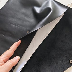 May include: A black leather envelope-style document holder with a smooth exterior finish and a textured suede interior. The folder features a metal snap button closure and contains several sheets of white paper inside the main compartment.