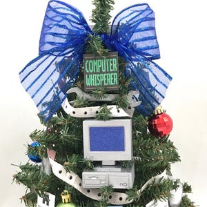 May include: A small artificial Christmas tree decorated with computer-themed ornaments, including a computer monitor, a wrench, and a sign that reads "Computer Whisperer". The tree is topped with a blue bow.