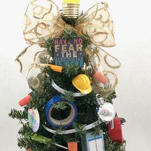May include: A small artificial Christmas tree decorated with electrician-themed ornaments, including a light bulb, a measuring tape, electrical tape, wire nuts, a hard hat, and a sign that reads "Have No Fear The Electrician Is Here".