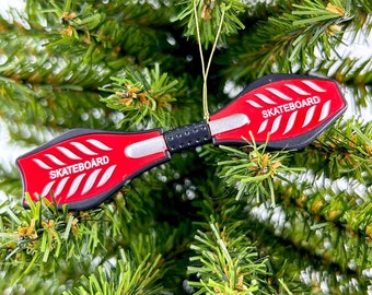 May include: Red and black skateboard ornament with the word "skateboard" printed on each side. The ornament is hanging from a green Christmas tree.
