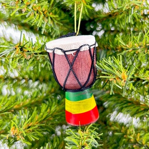 May include: A small wooden bongo drum ornament with a red, yellow, and green striped base. The drum is hanging from a green tree branch.