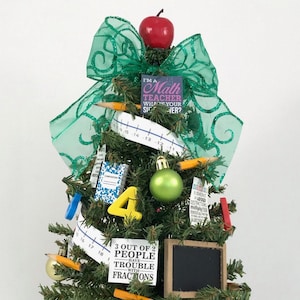 May include: A small artificial Christmas tree decorated with school-themed ornaments, including pencils, a ruler, a notebook, a chalkboard, and a green ornament. The tree is topped with a red apple and a green bow. The ornaments have text on them, including "I'm a Math Teacher, What's Your Super?" and "3 out of 2 People Have Trouble with Fractions."