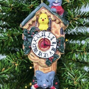 May include: A brown and tan cuckoo clock Christmas ornament with a yellow bird, a gray mouse on top, and a gray mouse on the bottom. The clock face has Roman numerals and a red hand.