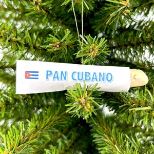 May include: A white paper tube ornament with the text "PAN CUBANO" in blue letters and a small Cuban flag printed on the left side. The ornament is hanging on a green artificial Christmas tree.