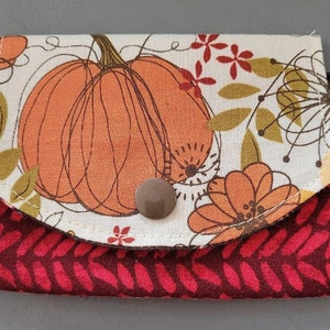 May include: A small, rectangular wallet with a fall theme. The top flap features a large orange pumpkin with brown outlines and floral accents on a cream background. The bottom section is a deep red with a repeating leaf pattern. A brown button secures the wallet.