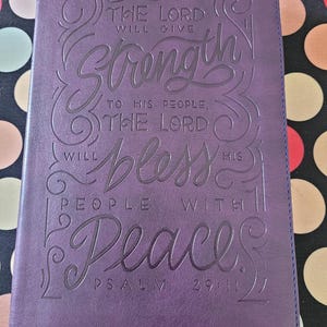Könnte beinhalten: Violettes Lederjournal mit geprägtem Text. Auf dem Einband steht "THE LORD WILL GIVE Strength TO HIS PEOPLE, THE LORD WILL BLESS HIS PEOPLE WITH Peace" und "PSALM 29:11". Mit roten und violetten Lesebändchen.