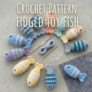 May include: A collection of small crocheted fish in various colors including blue, yellow, purple, and peach, arranged in a circle. The text CROCHET PATTERN FIDGET TOY FISH is visible. Includes a metal crochet hook, yarn, and plastic stitch markers.