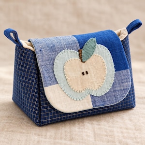 Puede incluir: Una pequeña bolsa de tela con cuerpo de cuadros azules y una solapa decorada con una aplicación de manzana superpuesta. El diseño de la manzana consta de piezas de tela azul claro y crema cosidas con un detalle de hoja verde.